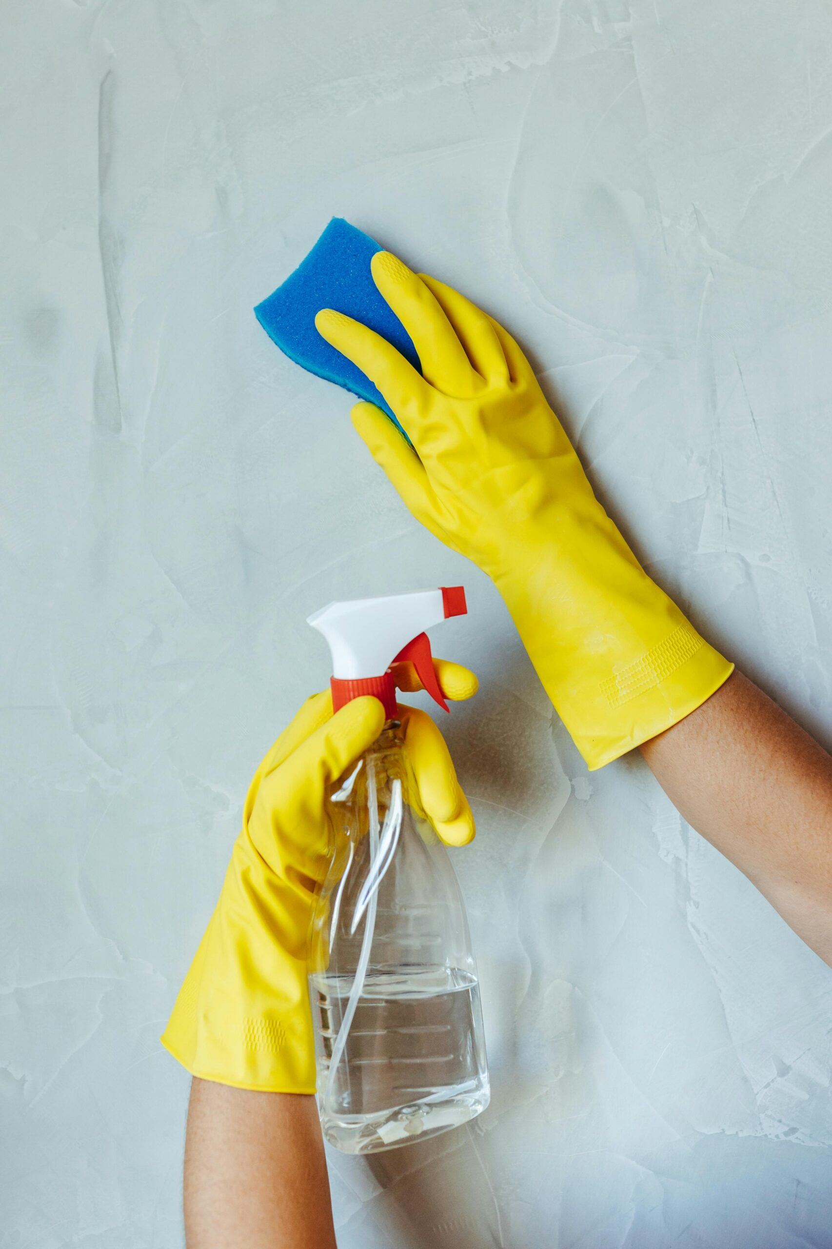 Hands in yellow gloves cleaning wall with sponge and spray bottle.